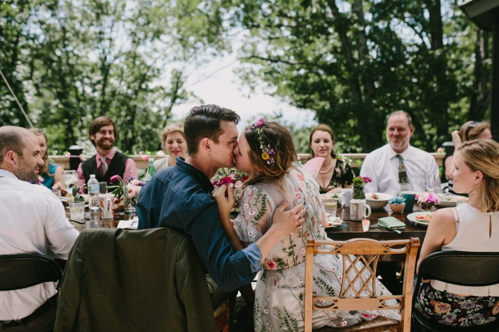 fairy bride in flower crown and floral dress and groom kissing at reception dinner with family surrounding