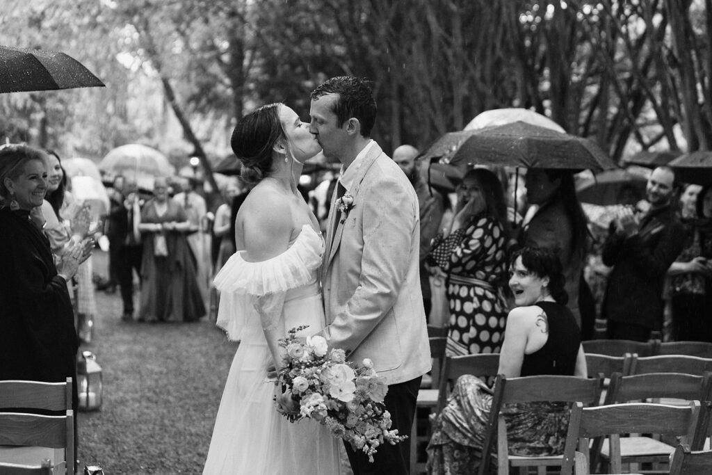 bride and groom kissing in the rain at the end of the aisle with smiling guests holding umbrellas in the background